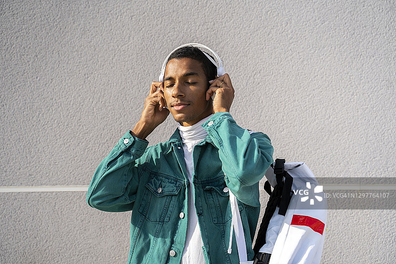 一个年轻人靠着灰色的墙，戴着耳机听音乐图片素材