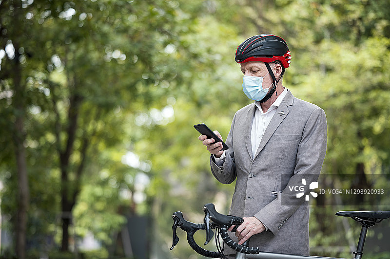 在城市工作的成熟商人用自行车同时使用手机。在疫情蔓延期间，他戴着口罩。图片素材