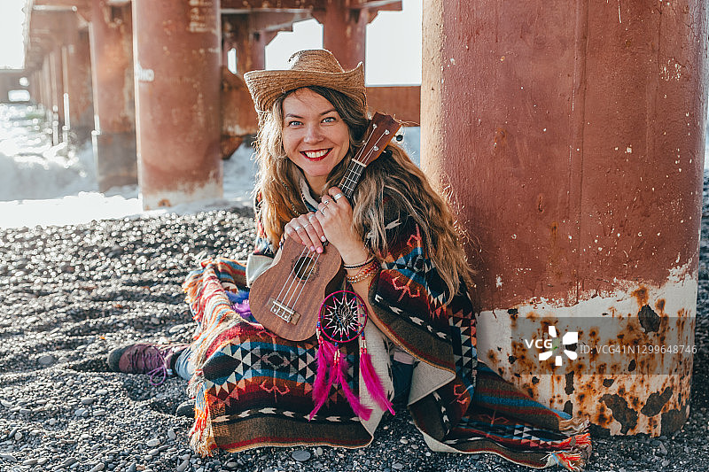一个年轻的嬉皮女人坐在码头附近的海滩上弹奏四弦琴。图片素材