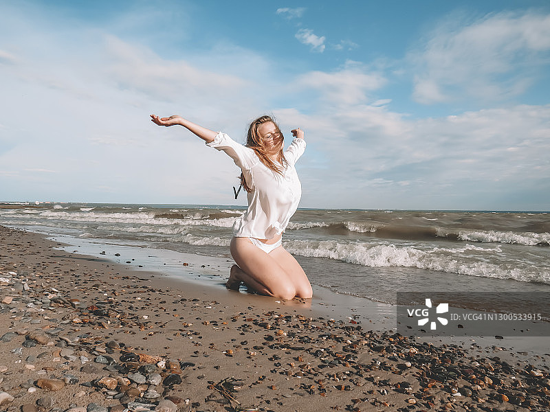 一位头发蓬乱的金发女郎，穿着白色衬衫和比基尼，一双晒黑了的腿修长在沙滩上，享受着海风和阳光的温暖图片素材