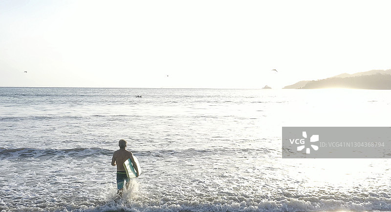 从鸟瞰图上看，一个成熟的男人扛着冲浪板进入柔和的海浪中图片素材