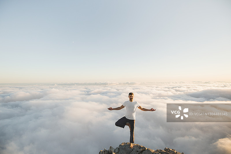 在早晨，一个人在山上独自练习哈他瑜伽(树式)，看到令人惊叹的低云图片素材