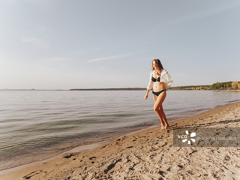 一位迷人的年轻女子穿着黑色比基尼和丝绸衬衫，晒黑的修长腿在日落时分沿着海边的水边奔跑图片素材