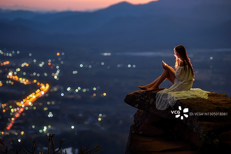 泰国，一名女子坐在城市上方的窗台上使用手机图片素材