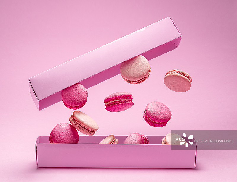 五颜六色的甜马卡龙或杏仁饼干，调味饼干漂浮在纸盒上方的空中。粉红色的背景。图片素材