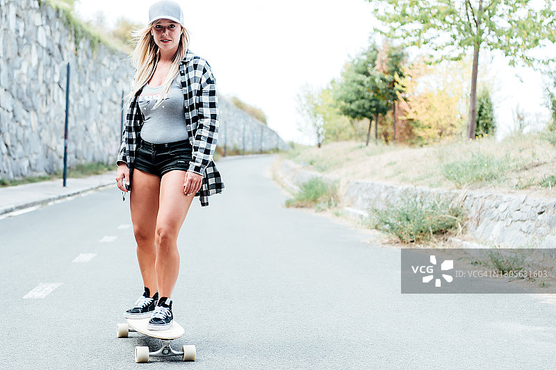 金发女人穿着黑色短裤，灰色帽子和格子衬衫滑下一个长滑板的道路。图片素材