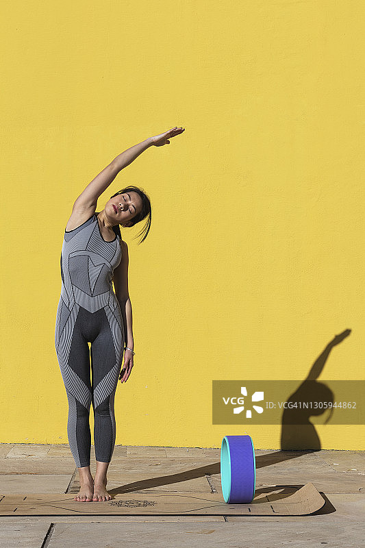 女人举起手臂，靠在黄色的墙上，靠在瑜伽轮上做运动图片素材