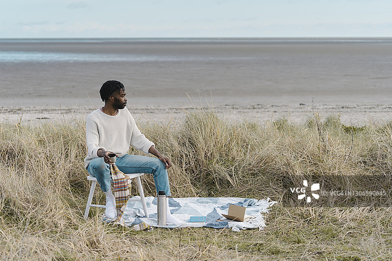 一个年轻人坐在沙滩上干枯的植物中间的凳子上喝茶图片素材