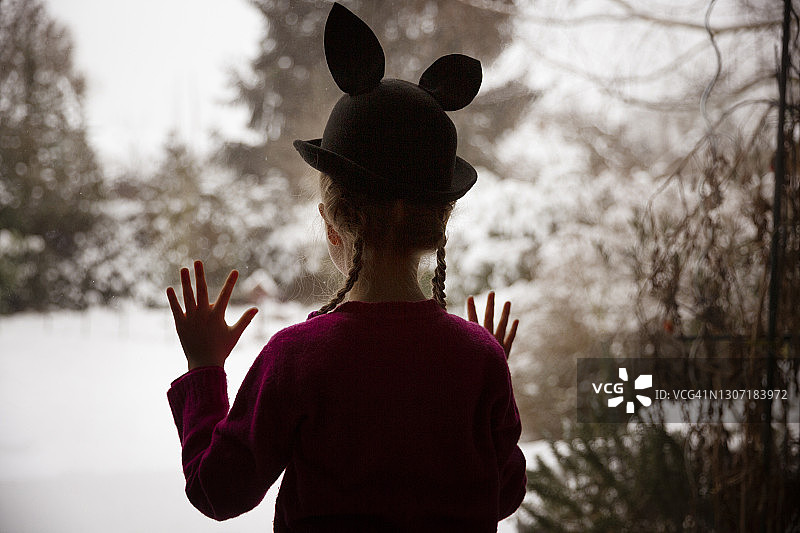 孩子戴着兔耳帽，望着窗外白雪覆盖的后院图片素材