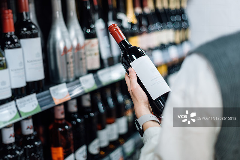 从肩膀上看，一个女人走过卖酒的过道，从超市的货架上挑选一瓶红酒图片素材