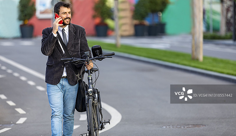一个穿着西装的年轻英俊的男人正在城市街道上骑着自行车和用手机聊天。图片素材