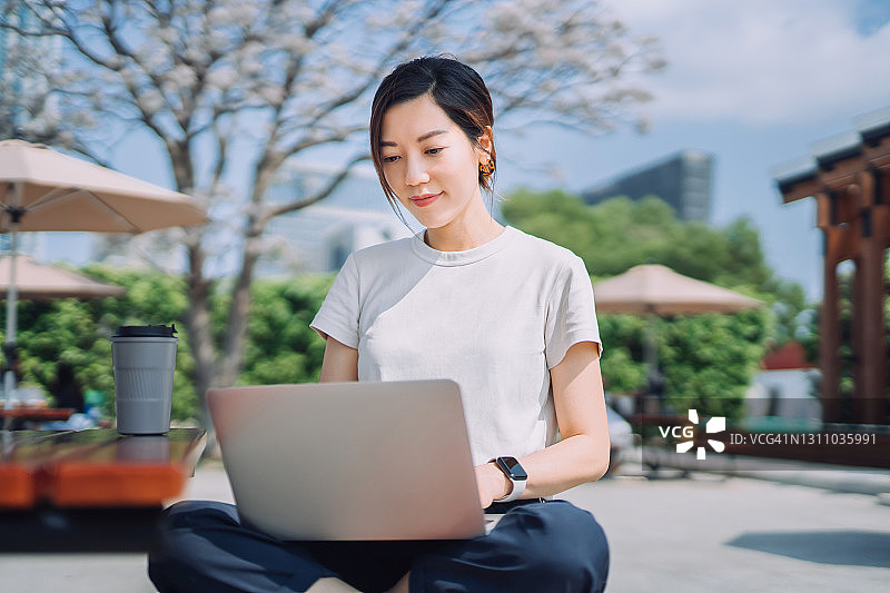 自信的年轻亚洲自由职业女商人在城市公园的户外用笔记本电脑工作，桌上放着可重复使用的咖啡杯。远程办公理念，灵活的生活方式图片素材
