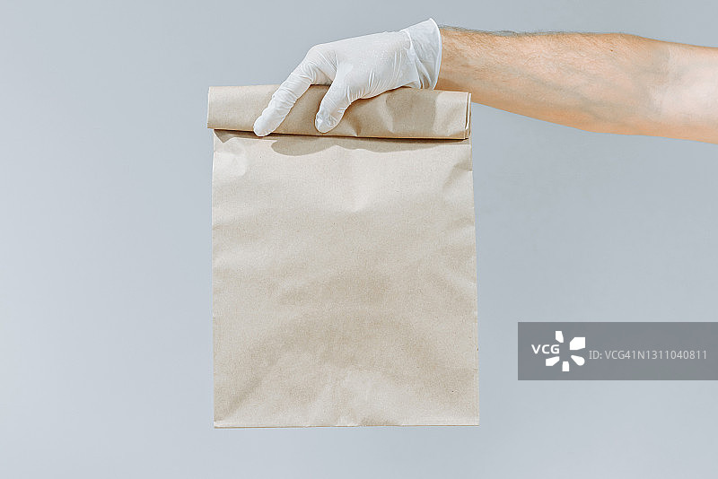 一个戴手套的男人的手拿着一个棕色的袋子或一个孤立的灰色背景的袋子。包装布局模板。送货服务的概念。副本的空间。快递图片素材