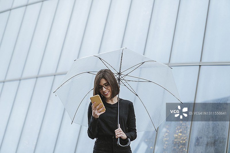 微笑的女人用手机，同时拿着雨伞对着玻璃建筑图片素材