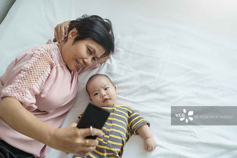 快乐的亚洲祖母和小男孩或孙子躺在床上自拍，用手机或智能手机打视频电话。图片素材