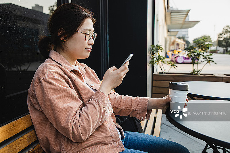 亚洲女性在咖啡馆使用电话，用可重复使用的咖啡杯喝咖啡图片素材