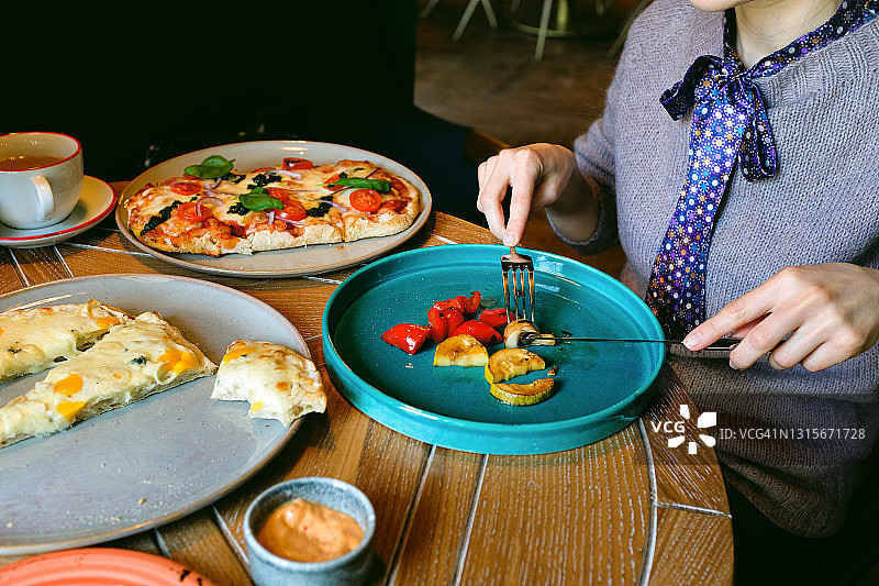 一位妇女将烤好的蔬菜切成片放在盘子里吃。披萨旁边的奶酪和披萨玛格丽特加罗勒叶和香蒜沙司，在咖啡馆或快餐店的桌子上。在刀叉的手中。意大利菜。素食。图片素材