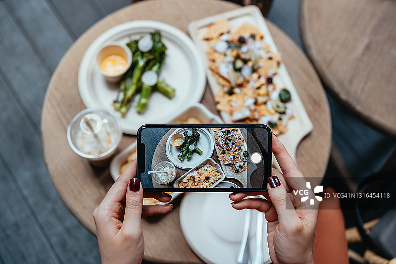 一名年轻女子在阳台上享受自家的外卖，在吃之前用智能手机拍下美味的食物。饮食生活习惯。相机先吃文化。日常生活中的科技图片素材