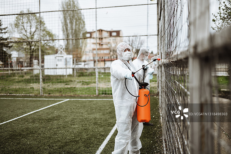 穿着防护服的现代医务工作者对室外足球场的围栏消毒图片素材