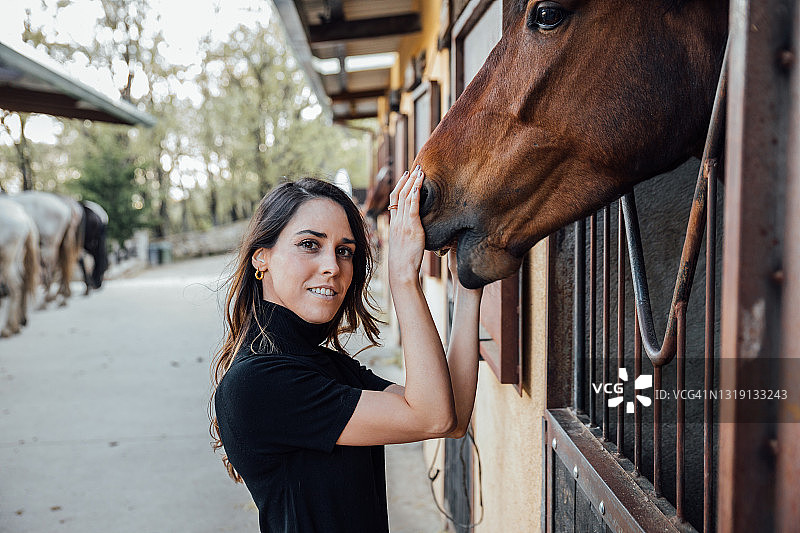 一个女人在看相机的时候抚摸着马厩里的棕色马的侧视图图片素材