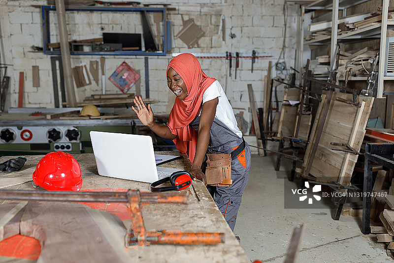 一名年轻的中东妇女戴着头巾，在大型木工车间里用笔记本电脑塑造木板。图片素材