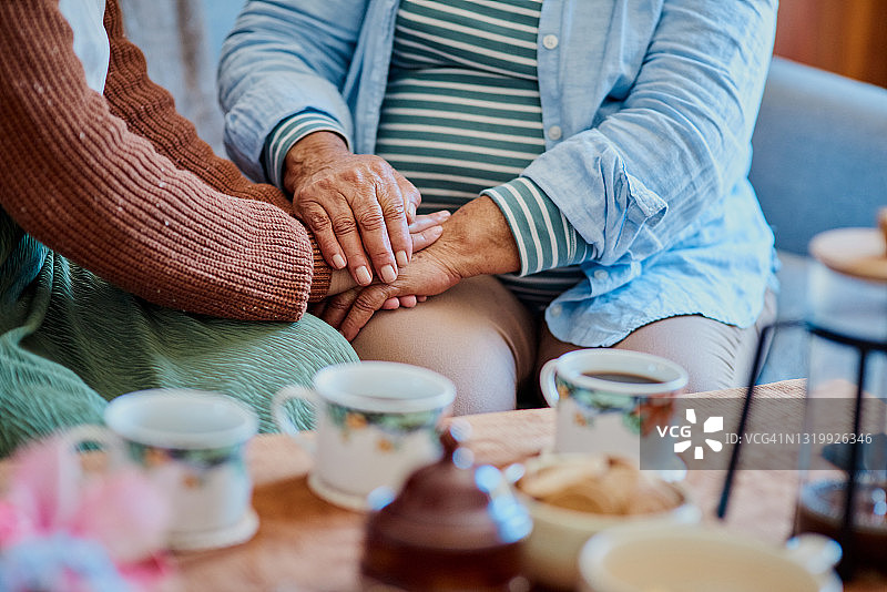 照片中，一名妇女和她年长的亲戚手牵着手，坐在家里的沙发上喝咖啡图片素材