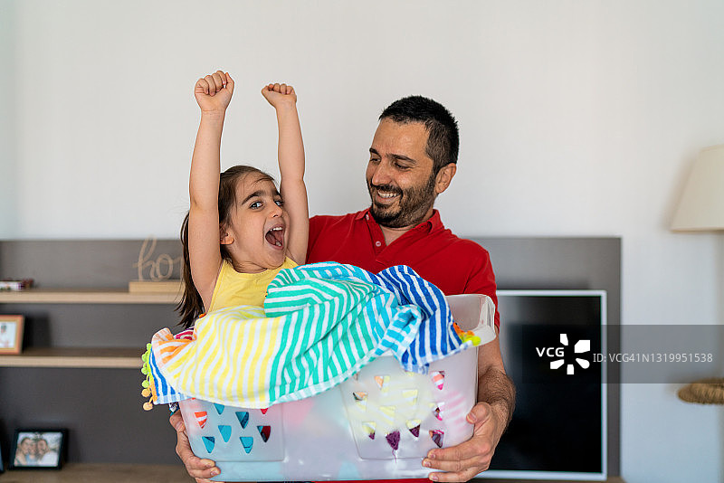 年轻的父亲试图把衣服放在洗衣篮在家里，与他的女儿坐在洗衣篮里玩图片素材
