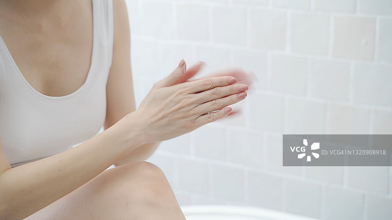 亚洲妇女短发，在白色浴室附近的浴缸，在家庭工作室装饰一些绿色植物涂霜乳液的手，腿和手臂。白色霜摩擦治疗皮肤状况。擦洗图片素材