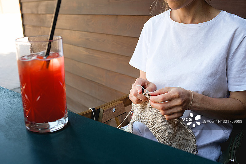 一名年轻女子坐在露天的木椅或长凳上，用编织针编织一件时髦的现代时尚开衫、夹克或毛衣。冷却不含酒精的草莓饮料或鸡尾酒。一种爱好的概念，世界编织日公开。图片素材