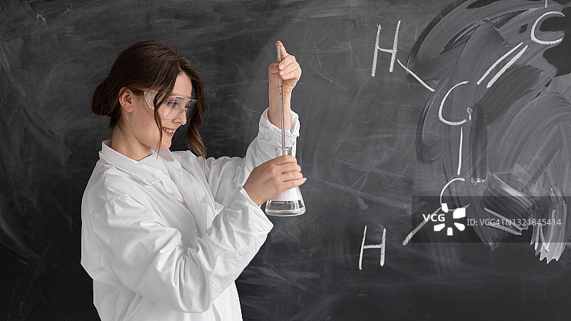一位化学老师站在学校里，黑板的背景是化学公式。白色的病号服和护目镜他手里拿着一个烧瓶，做了一个实验。概念回到学校。图片素材