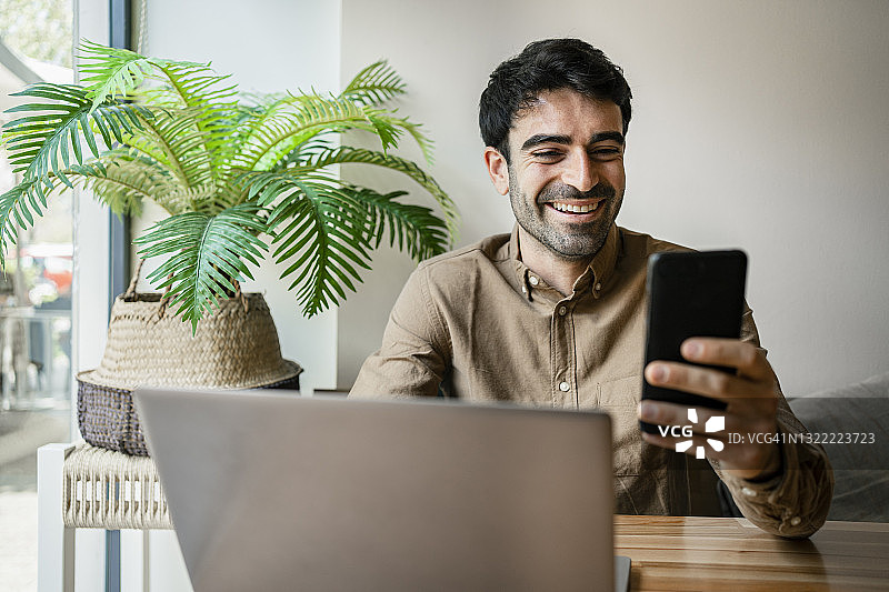 微笑的男性自由职业者使用手机，而坐在咖啡馆笔记本电脑图片素材