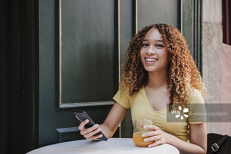 微笑的女人拿着橙汁，坐在路边咖啡馆的智能手机图片素材