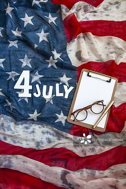 俯视图“7月4日”由数字和白色木制字母、空白剪贴板、眼镜和美国国旗上的金色铅笔组成。7月4日，独立日。图片素材