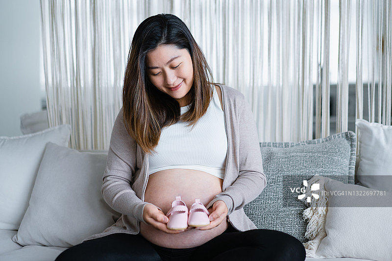 微笑的年轻亚洲孕妇坐在舒适的家里的沙发上，拿着一双粉红色的婴儿鞋靠在她的肚子上。孕妇。期待新的生活理念图片素材