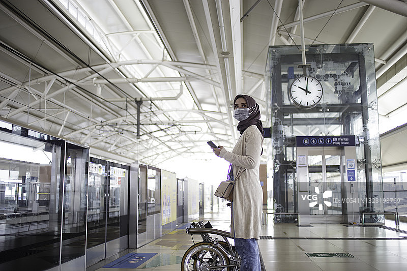亚洲年轻女子戴着头巾，骑着自行车，在站台上打电话，等捷运图片素材