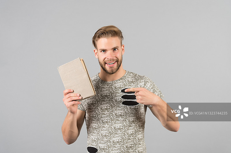 一个留着胡子，穿着时髦衬衫的男人指着一本书。男人微笑着推荐书，灰色背景。文献推荐的概念。一位现代时尚作家出版了一本书图片素材