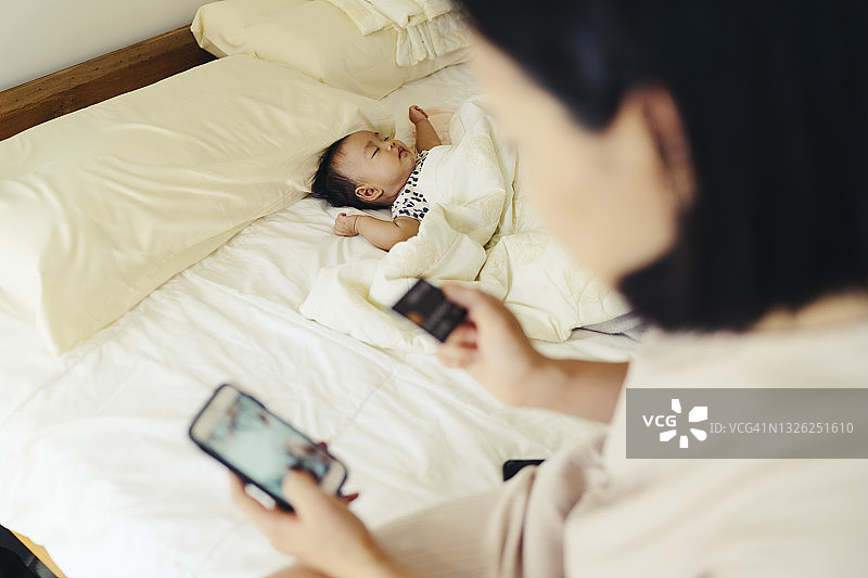 年轻的亚洲母亲用手机和批准的信用卡购买东西，而女儿睡在床上图片素材