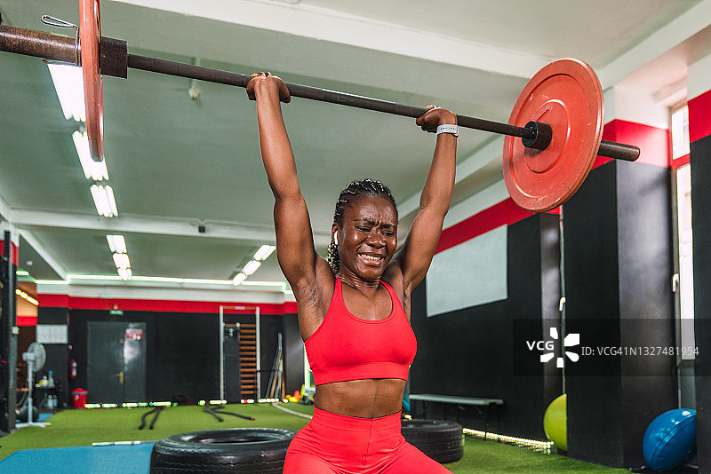 强壮的黑人运动员在体育馆做杠铃和举重练习，以加强肩膀和肱二头肌，显示出很多努力和红色运动服图片素材