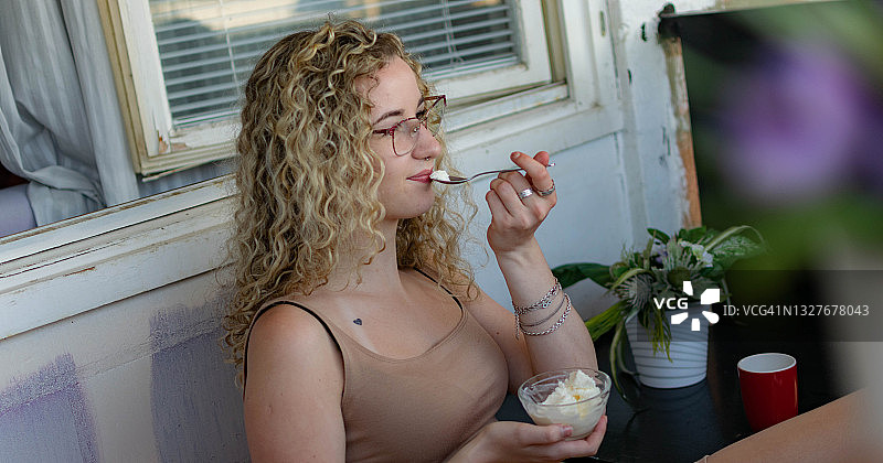 年轻的卷发女子坐在家里，享受着吃冰淇淋，享受着新鲜空气，吃着冰激凌在她的阳台上，同时使用着笔记本电脑。图片素材