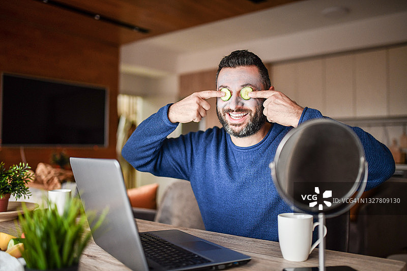 微笑的男性一边用笔记本电脑和咖啡放松，一边在眼睛上涂黄瓜作为面膜图片素材