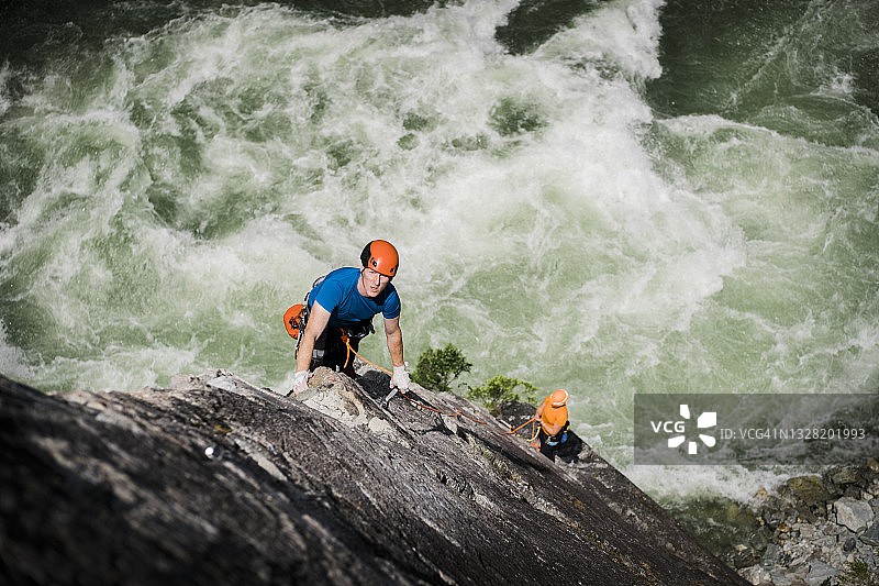 一名男性攀岩者在一座高耸于河流之上的戏剧性岩壁上，将攀爬绳系在锚上图片素材