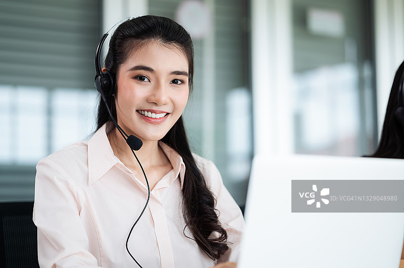 亚洲呼叫中心的员工带着耳机和麦克风。在柜台和电脑前为顾客服务时要微笑。服务理念与咨询。通信的概念。图片素材