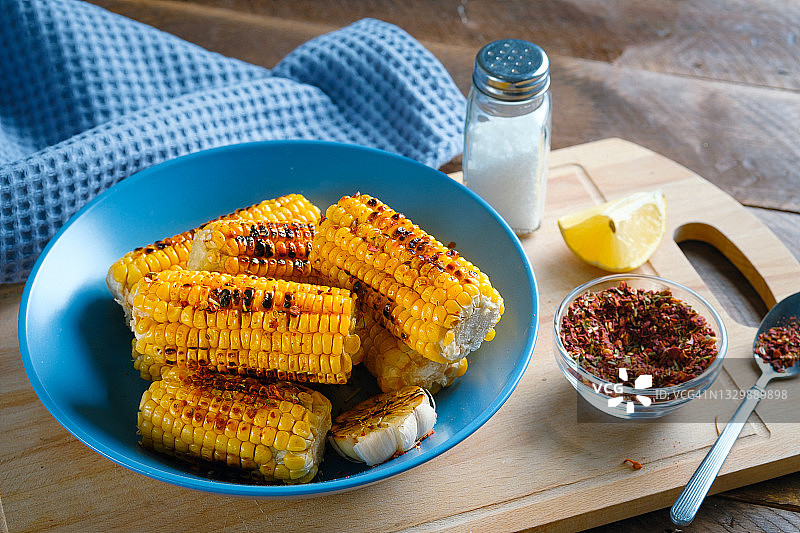 黄色的玉米，烤过，撒上香料，放在一个明亮的陶瓷盘子上，背景是木制的厨房桌子。家庭自制食物的概念。素食和纯素食食品和饮食。图片素材