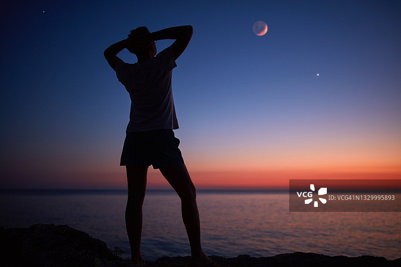 一个人的剪影看着月亮和星星越过海洋的地平线。图片素材