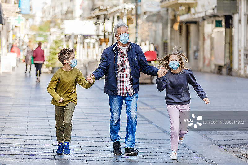 一位戴着防护口罩的老人带着孙子和孙女走在城市的街道上。图片素材