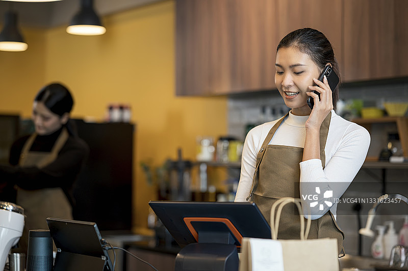 一位亚洲女咖啡店老板通过智能手机与顾客确认订单，然后将订单发送给咖啡师制作咖啡。食品和饮料订购系统，外卖食品，和小企业主。图片素材