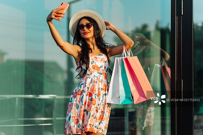 美丽时尚的年轻女子拎着购物袋，站在城市街道上用智能手机自拍图片素材