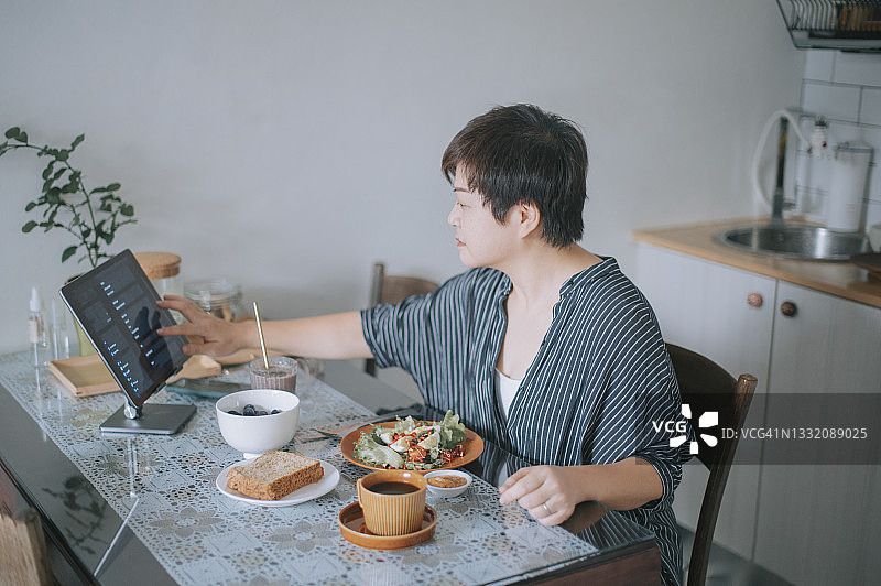 一名亚洲华人女子在餐桌上吃着健康的沙拉早餐，一边用数字平板电脑滚动音乐列表图片素材