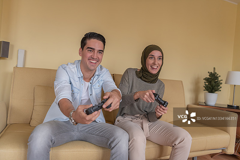 高兴的年轻人和他的穆斯林女友戴着头巾在家里玩电子游戏。图片素材