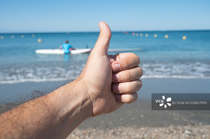 竖起大拇指。一个男人在海边的沙滩上做了一个“可以”的手势。背景是一个人在水中学习冲浪板。图片素材